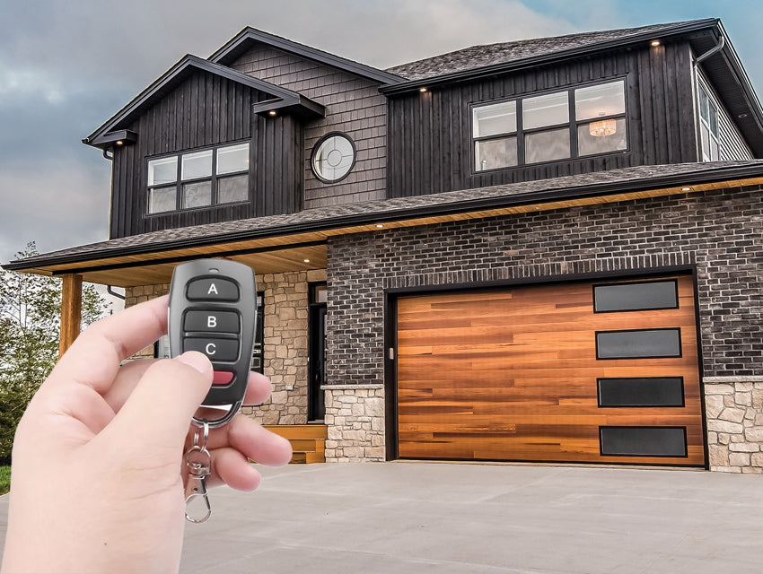 Modern two-story house with black siding, stone accents, and a distinctive wooden garage door, featuring a hand holding a remote control.